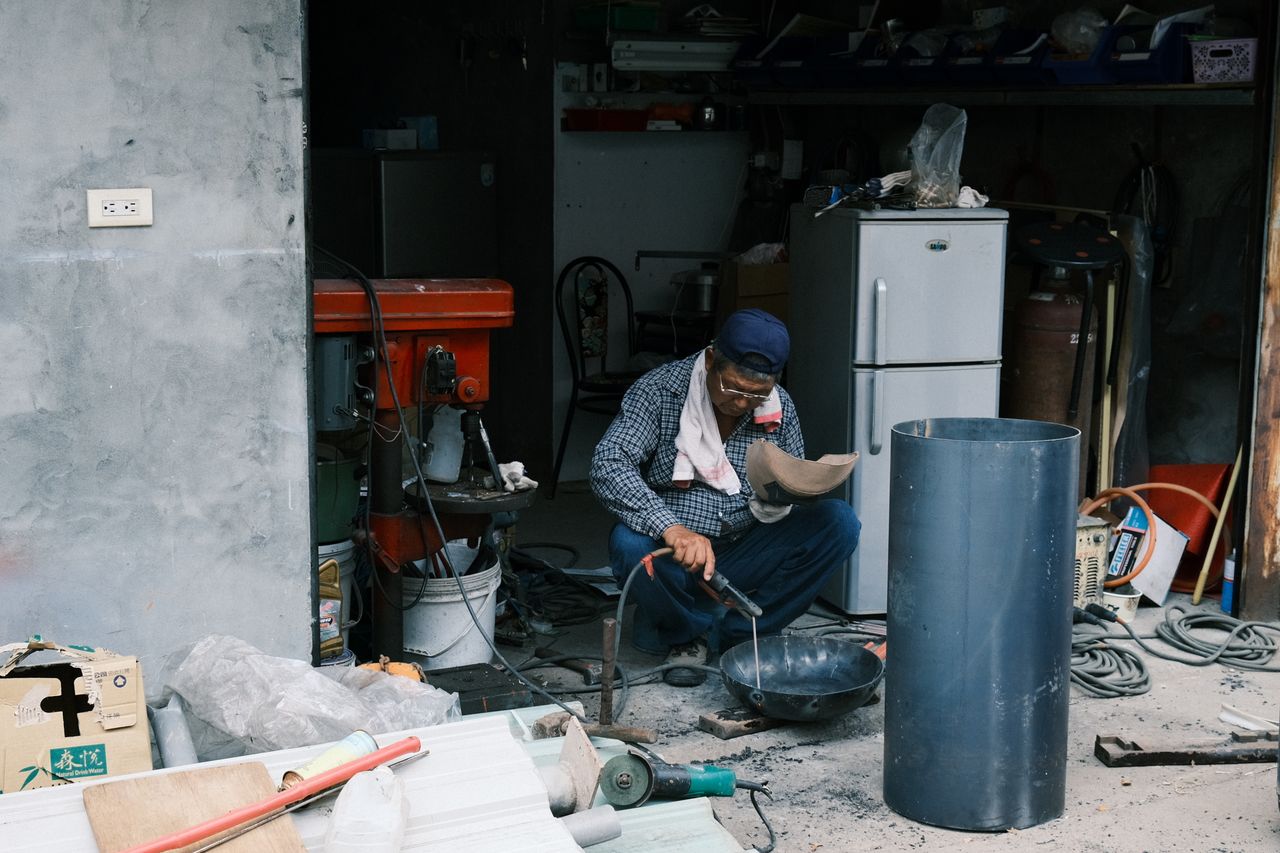 An adult man welding in a garage in Taiwan, surrounded by tools and equipment.
